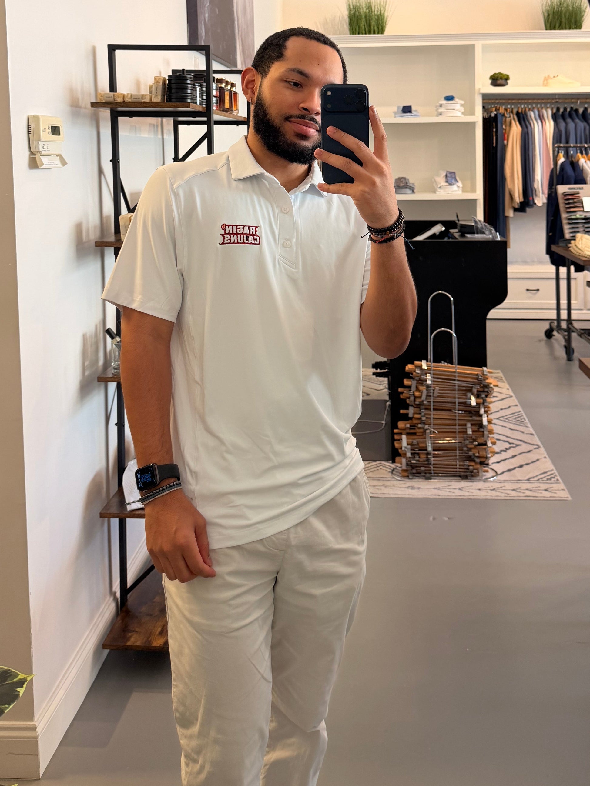 A man takes a mirror selfie in a clothing boutique, wearing a University of Louisiana-Lafayette Ragin Cajuns white polo shirt with a Ragin Cajuns red embroidered logo on the chest and white pants. The boutique has a modern interior with a large arched mirror, clothing racks, and shelves.