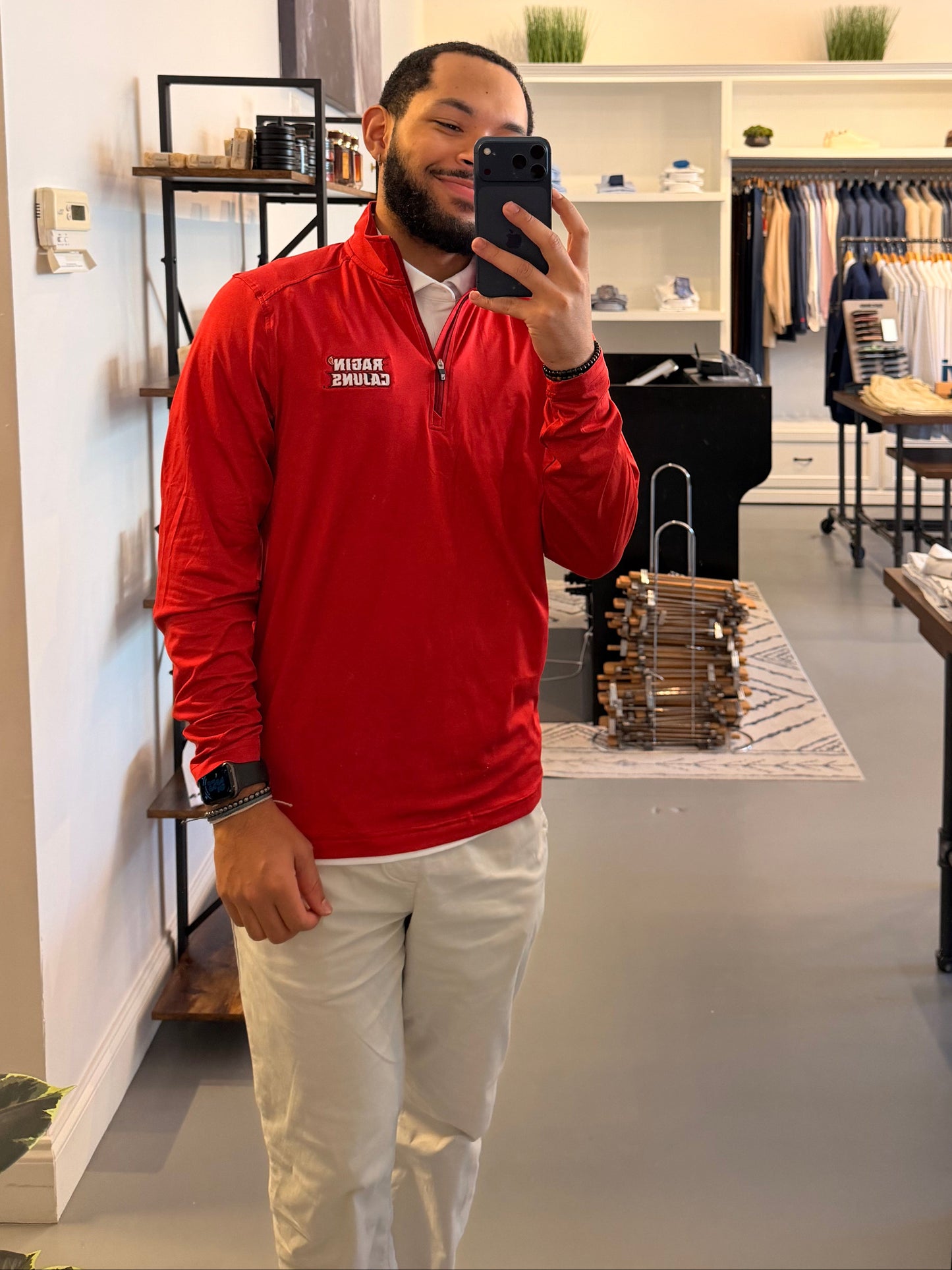 A man takes a mirror selfie in a clothing boutique, wearing a Cutter & Buck University of Louisiana-Lafayette red long-sleeve quarter-zip pullover with Ragin Cajuns embroidered on the left chest, paired with off-white pants.