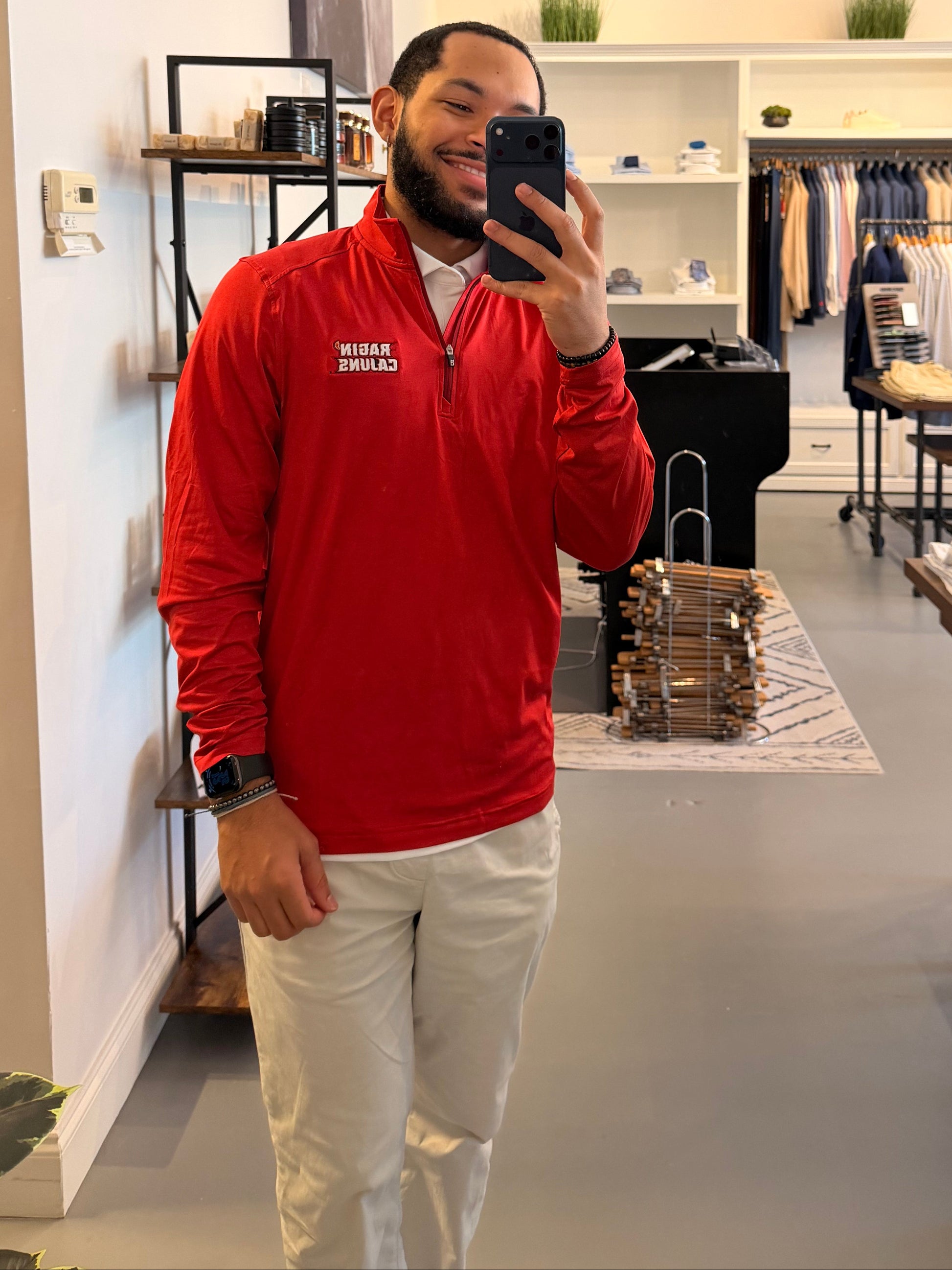 A man takes a mirror selfie in a clothing boutique, wearing a Cutter & Buck University of Louisiana-Lafayette red long-sleeve quarter-zip pullover with Ragin Cajuns embroidered on the left chest, paired with off-white pants.