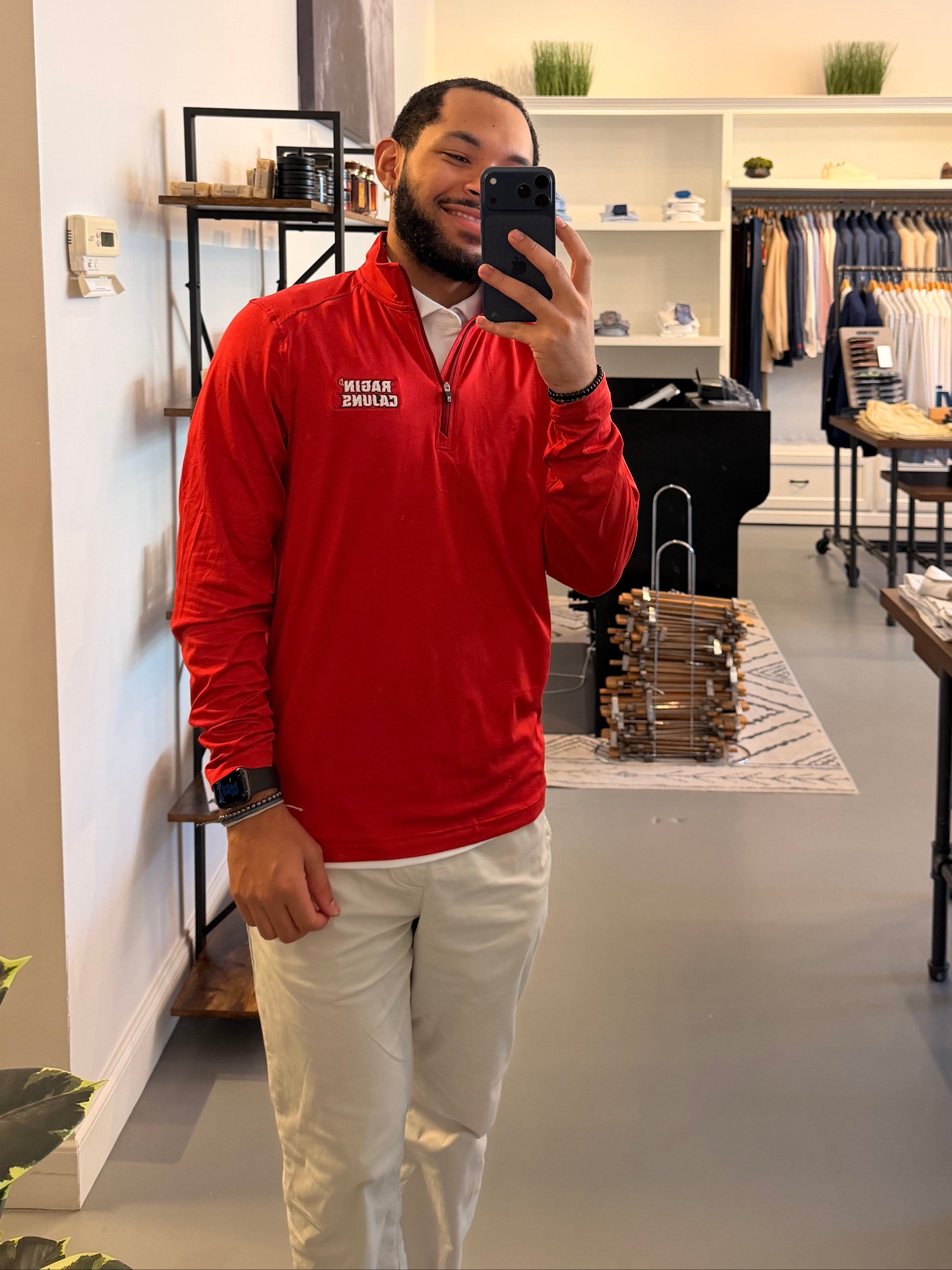 A man takes a mirror selfie in a clothing boutique, wearing a Cutter & Buck University of Louisiana-Lafayette red long-sleeve quarter-zip pullover with Ragin Cajuns embroidered on the left chest, paired with off-white pants.