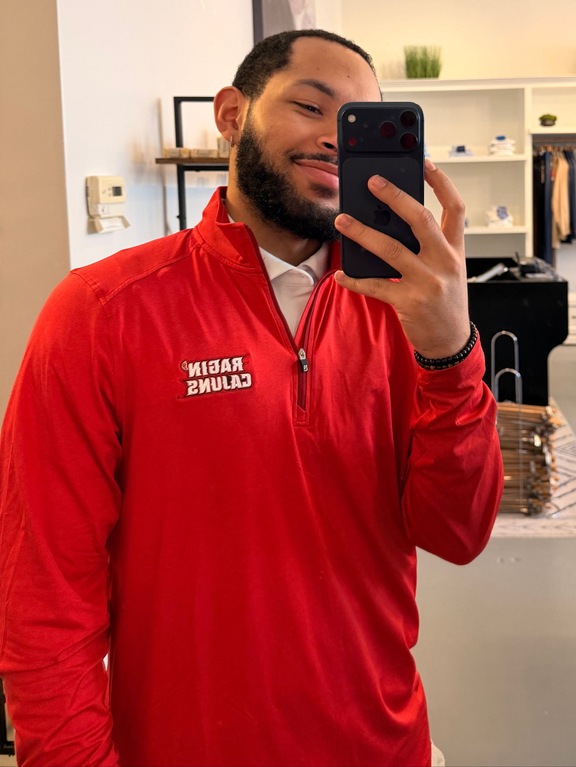 A man takes a mirror selfie in a clothing boutique, wearing a Cutter & Buck University of Louisiana-Lafayette red long-sleeve quarter-zip pullover with Ragin Cajuns embroidered on the left chest, paired with off-white pants.